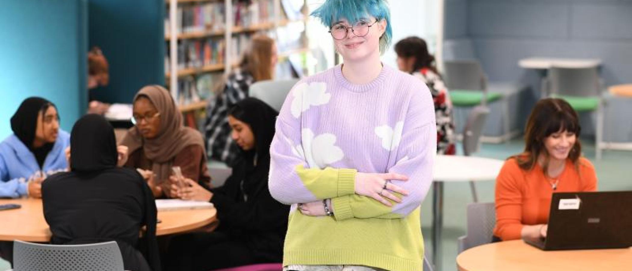 A student with blue hair and crossed arms standing and smiling at the camera, with students working and chatting on tables in the background