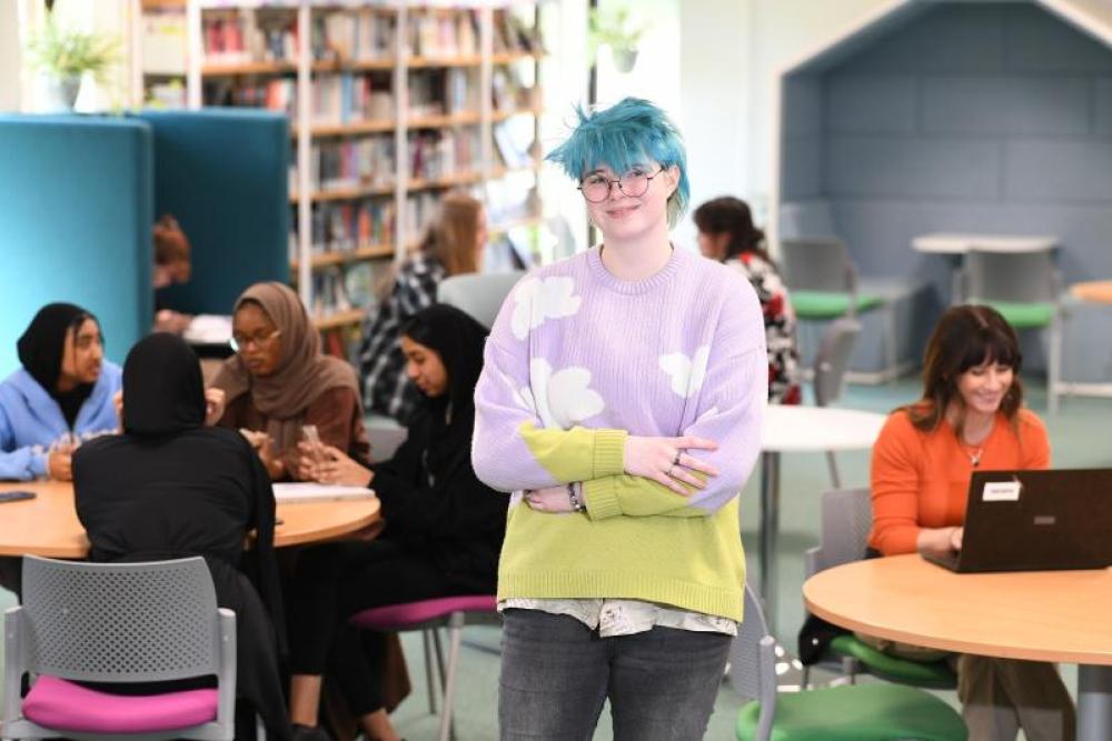 A student with blue hair and crossed arms standing and smiling at the camera, with students working and chatting on tables in the background
