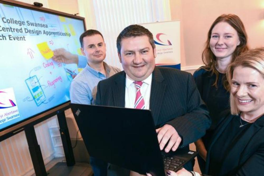 Bruce Fellowes, Head of GCS Training, is pictured holding a laptop in front of a screen which says Gower College Swansea User Centred Design Apprenticeship Launch Event. Bruce is pictured alongside James Holloway, Digital Trainer and Assessor at Gower College Swansea, Lauren Power, Product Manager at Digital Public Services Wales, and Sarah Floyd, Apprentice at Digital Public Services Wales.