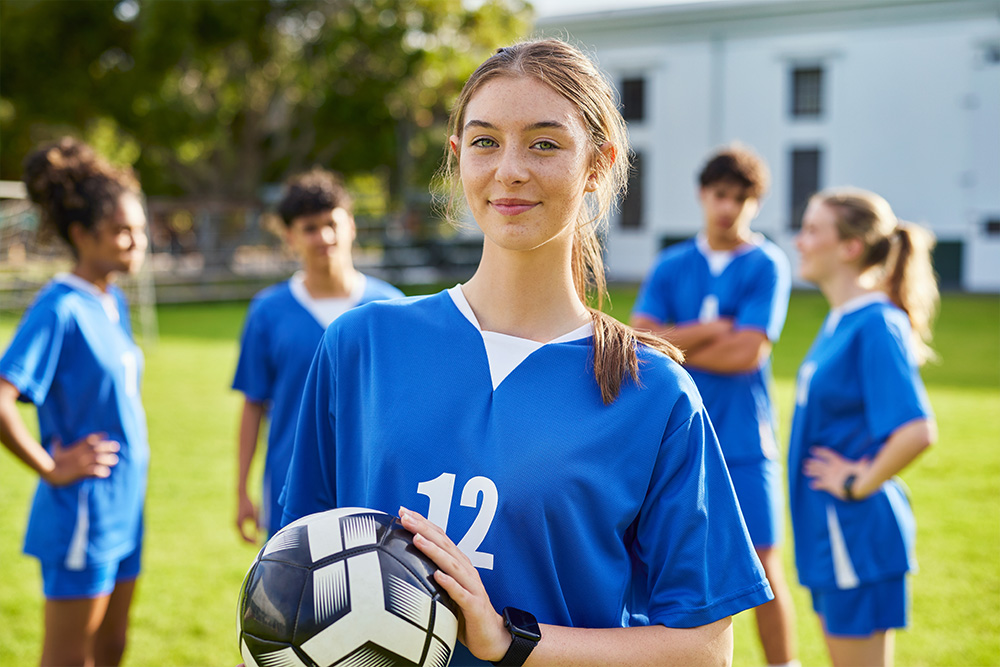 Teenager holds a football as she looks at the camera, with a group of other players behind her