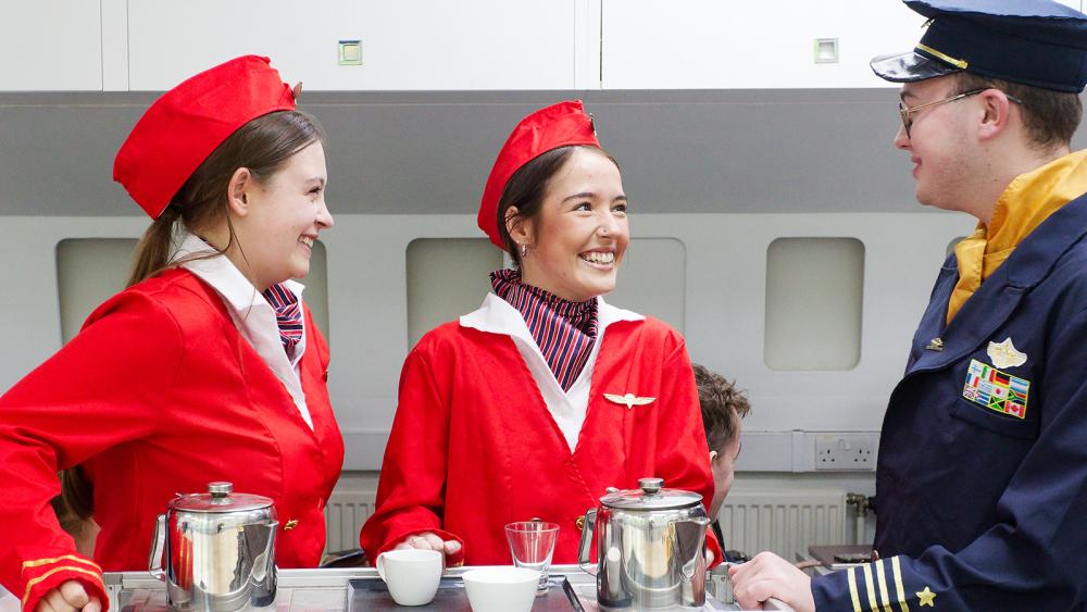 Two travel students in cabin crew uniform and one student in a pilot uniform practicing in the mock-up of a plane