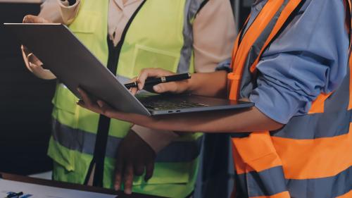 Two people in hi-vis vests looking at a laptop. One holds the laptop and has a pen in the other hand. Desk has forecasts and building documentation. There is a small model of a wind turbine also.