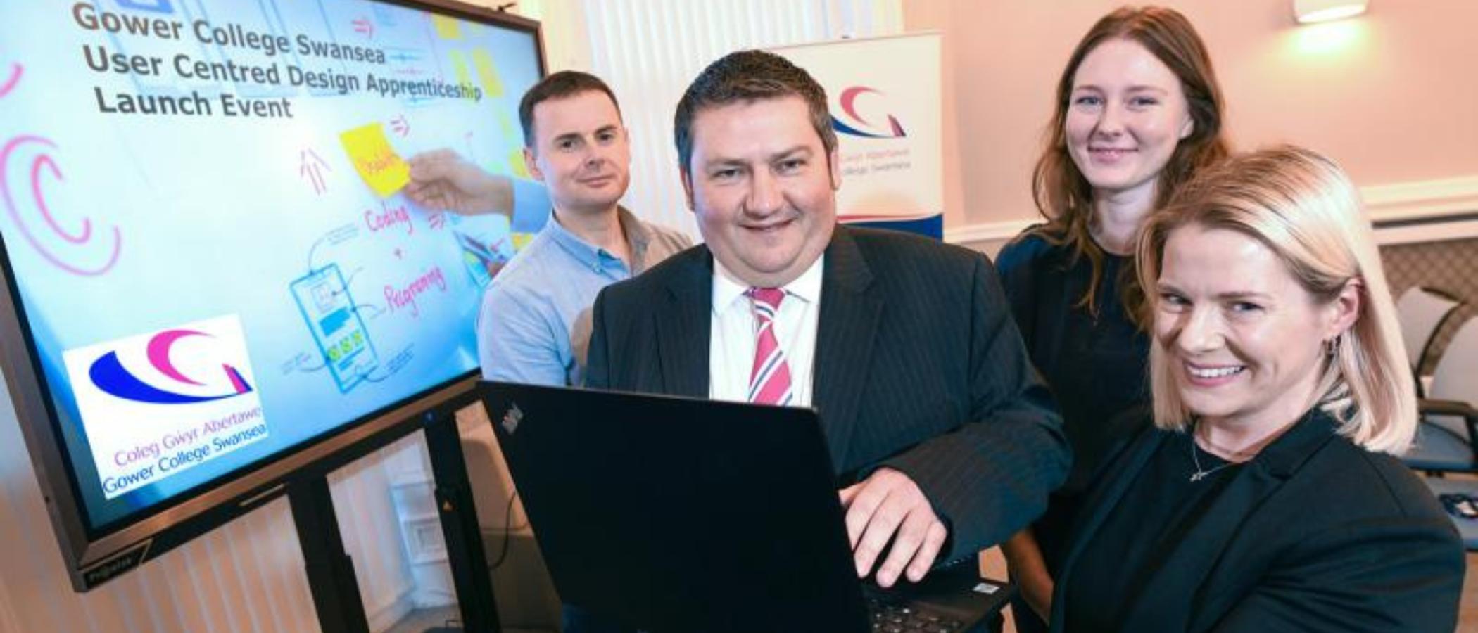 Bruce Fellowes, Head of GCS Training, is pictured holding a laptop in front of a screen which says Gower College Swansea User Centred Design Apprenticeship Launch Event. Bruce is pictured alongside James Holloway, Digital Trainer and Assessor at Gower College Swansea, Lauren Power, Product Manager at Digital Public Services Wales, and Sarah Floyd, Apprentice at Digital Public Services Wales.