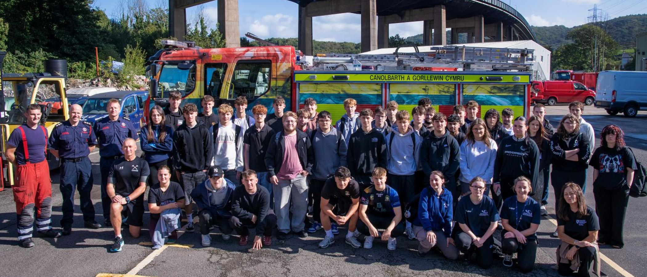 Large group of people standing next to fire engine, bridge in background and blue sky